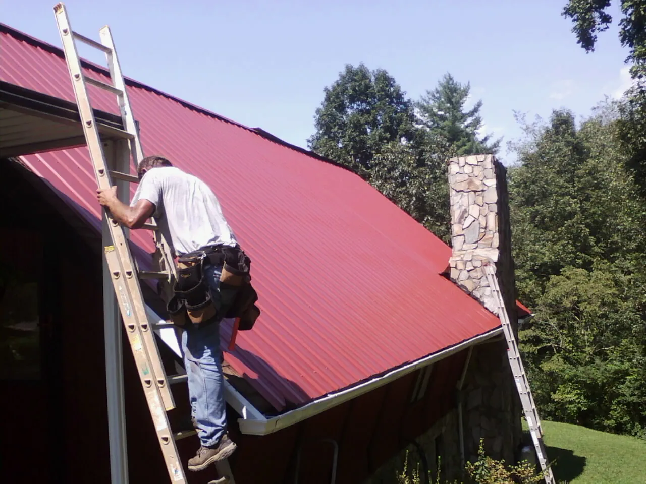 Roofer on a ladder beside a red metal roof and stone chimney — Soffit & Fascia Repair by Roofing Brothers in Grangeville